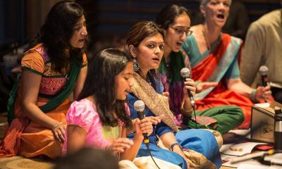 Girl speaking on microphone is surrounded by women wearing traditional clothing
