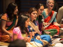 Girl speaking on microphone is surrounded by women wearing traditional clothing