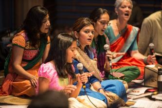 Girl speaking on microphone is surrounded by women wearing traditional clothing