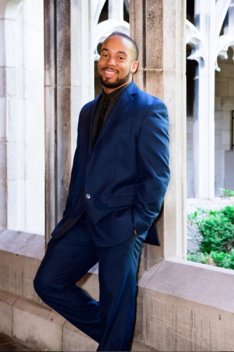 Man in blue suit leaning against a column smiling